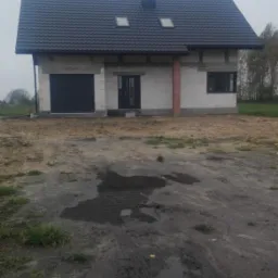 Surowy stan domu murowanego z ciemną dachówką i dwoma oknami dachowymi, widoczne niedokończone otoczenie i błotniste podłoże.