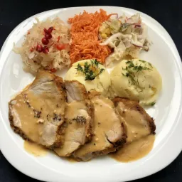 Porcja obiadowa na białym talerzu: kotlet schabowy w sosie musztardowym, ziemniaki puree z koperkiem, surówka z kiszonej kapusty z granatem, marchewka tarta i sałatka colesław.