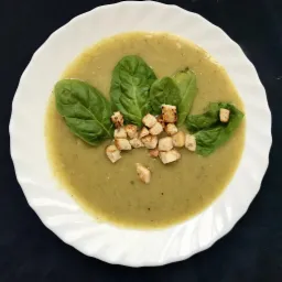 Kremowa zupa z zielonych warzyw na białym talerzu, udekorowana świeżymi liśćmi szpinaku i grzankami z chleba.