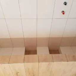 Układanie białych, prostokątnych płytek na suficie z widocznymi otworami na instalacje i drewnianym wykończeniem ściany w dolnej części kadru.