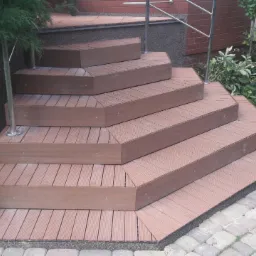 Zewnętrzne schody z desek kompozytowych w kolorze brązowym, z metalową barierką, otoczone zielenią i brukowaną ścieżką.