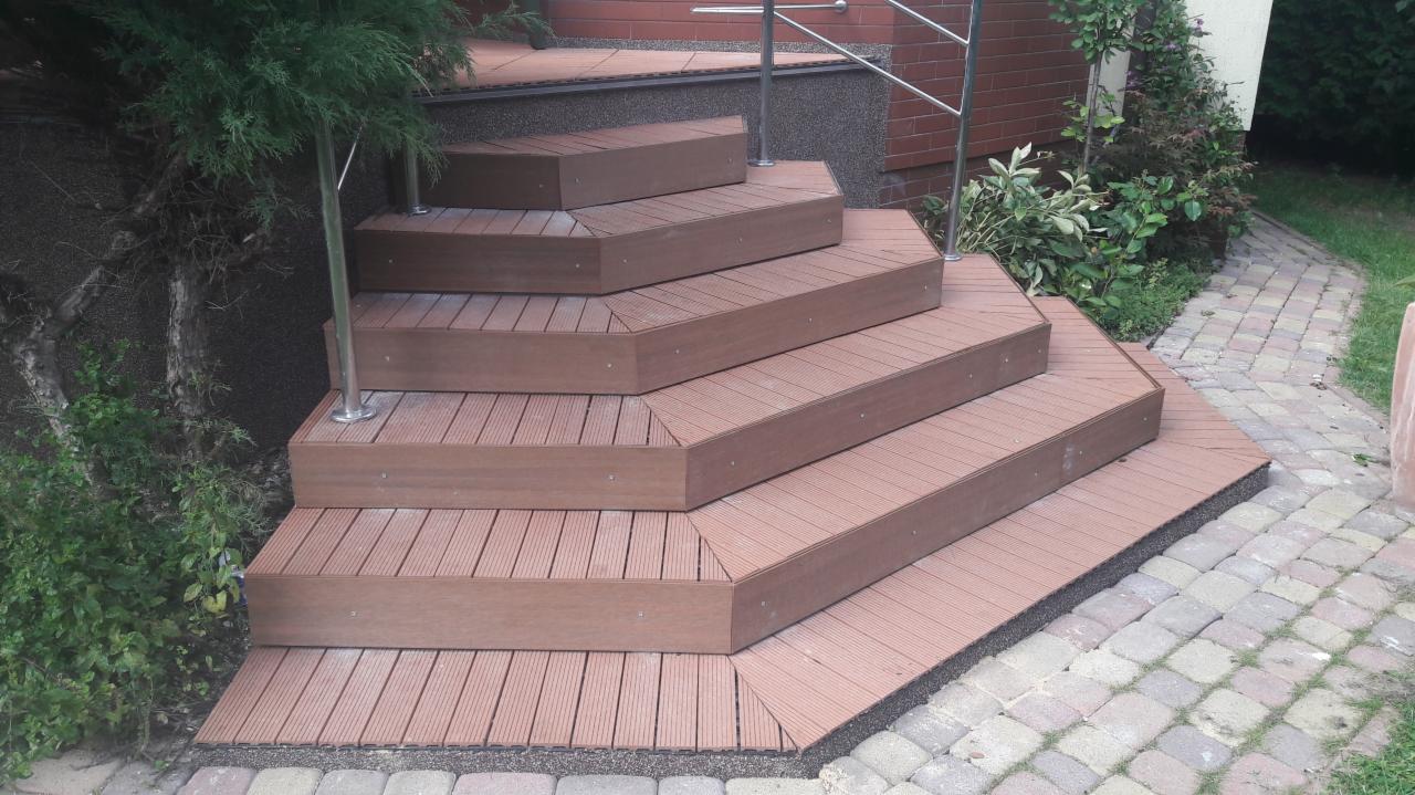 Zewnętrzne schody z desek kompozytowych w kolorze brązowym, z metalową barierką, otoczone zielenią i brukowaną ścieżką.