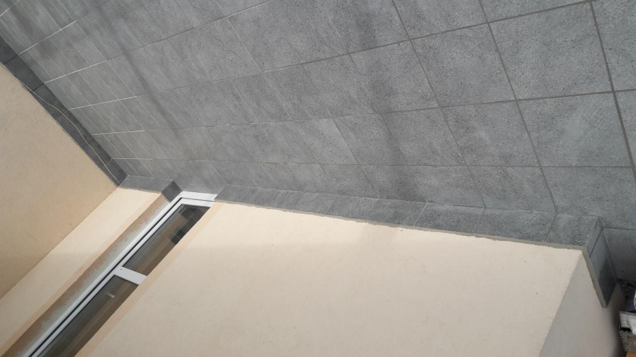 Szare, kwadratowe płytki ceramiczne ułożone na suficie, z widocznym fragmentem ściany i oknem w dolnej części kadru.