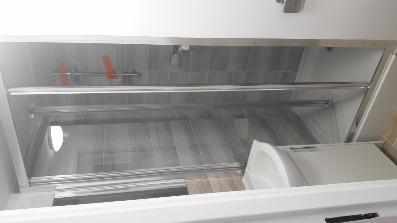 Szklana kabina prysznicowa z przesuwnymi drzwiami i szarymi płytkami w tle, widok z góry z perspektywy wanny.