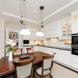 CITY CREATE BARTOSZ WANOT - Elegancka kuchnia z białymi szafkami na wymiar, ciemnym blatem i drewnianym stołem jadalnym z czterema krzesłami, oświetlona wiszącymi lampami z metalowymi elementami.