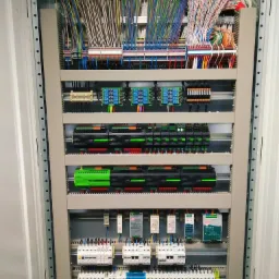 Rozdzielnica elektryczna z licznymi przewodami i modułami sterującymi, zainstalowana w Pozezdrzu. Widoczne kable uporządkowane w wiązki, zasilacze impulsowe i bezpieczniki.