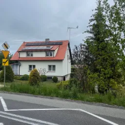 Dom z panelami słonecznymi i małą turbiną wiatrową na dachu, widziany z ulicy z znakami drogowymi. Biała elewacja i czerwona dachówka.