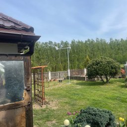 CITY CREATE BARTOSZ WANOT - Ogród z małą turbiną wiatrową na tle lasu, widoczna zardzewiała konstrukcja i fragment dachu budynku. Energia odnawialna w krajobrazie.