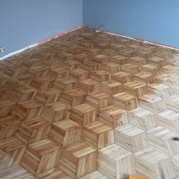 CITY CREATE BARTOSZ WANOT - Ukończona podłoga drewniana z wzorem 3D ułożona w rogu pomieszczenia. Widoczne narzędzie i puszka farby. Nietypowy kąt ujęcia podkreśla perspektywę podłogi.