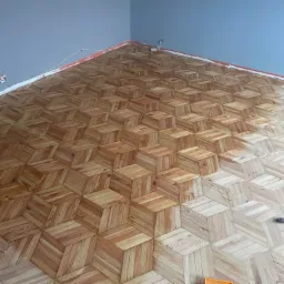 Ukończona podłoga drewniana z wzorem 3D ułożona w rogu pomieszczenia. Widoczne narzędzie i puszka farby. Nietypowy kąt ujęcia podkreśla perspektywę podłogi.