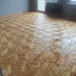 CITY CREATE BARTOSZ WANOT - Ukończona podłoga parkietowa z wzorem 3D w jasnym odcieniu drewna, świeżo po lakierowaniu, widok na całe pomieszczenie z grzejnikiem i oknem.