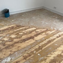 CITY CREATE BARTOSZ WANOT - Częściowo cyklinowana podłoga drewniana w jasnym pomieszczeniu. Widoczne deski z resztkami starej powłoki i pyłu. W rogu niebieski pojemnik z pokrywką.