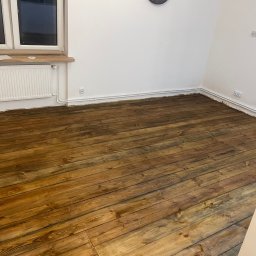 CITY CREATE BARTOSZ WANOT - Lakierowana, drewniana podłoga po cyklinowaniu w jasnym pomieszczeniu z białymi ścianami, grzejnikiem i oknem. Widoczne ślady po remoncie przy listwach przypodłogowych.