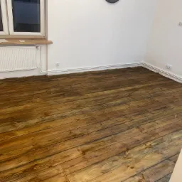 Lakierowana, drewniana podłoga po cyklinowaniu w jasnym pomieszczeniu z białymi ścianami, grzejnikiem i oknem. Widoczne ślady po remoncie przy listwach przypodłogowych.