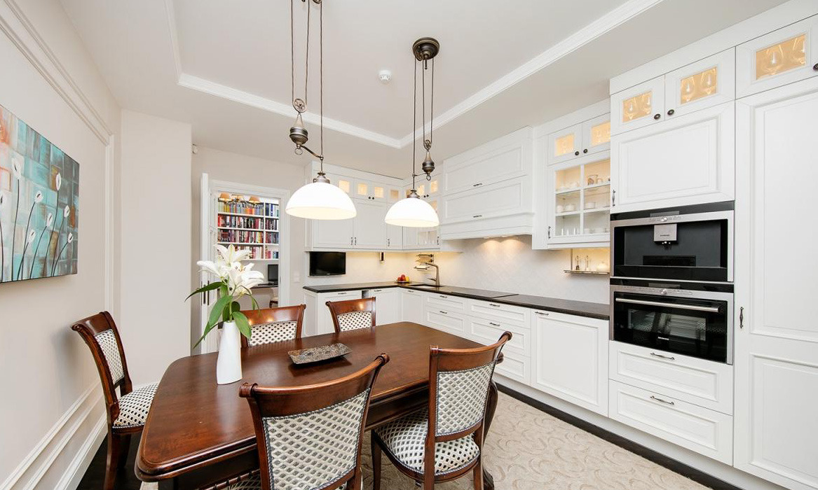 Elegancka kuchnia z białymi szafkami na wymiar, ciemnym blatem i drewnianym stołem jadalnym z czterema krzesłami, oświetlona wiszącymi lampami z metalowymi elementami.