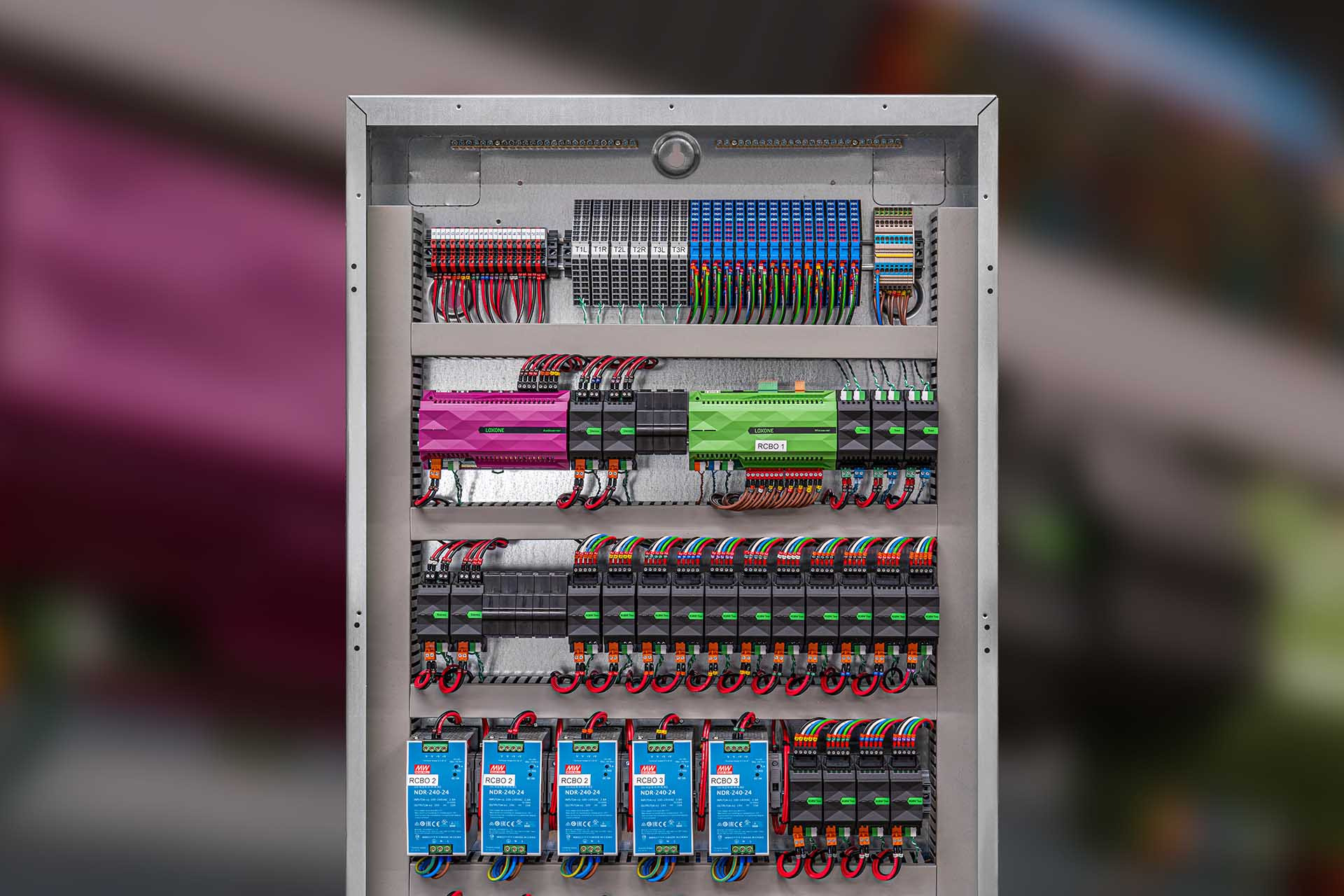 Rozdzielnica elektryczna z modułami sterującymi Loxone w kolorach różowym i zielonym, liczne przewody i złączki, widok z przodu na kompletną instalację w metalowej obudowie.