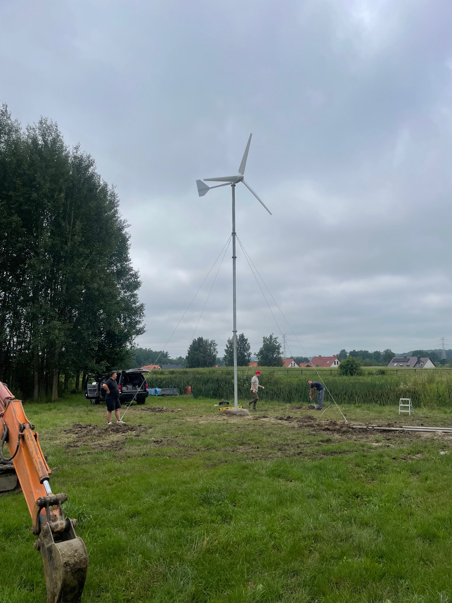 Montaż turbiny wiatrowej na łące. Widoczni pracownicy, wykopana ziemia i sprzęt budowlany. W tle zabudowania mieszkalne i pochmurne niebo.
