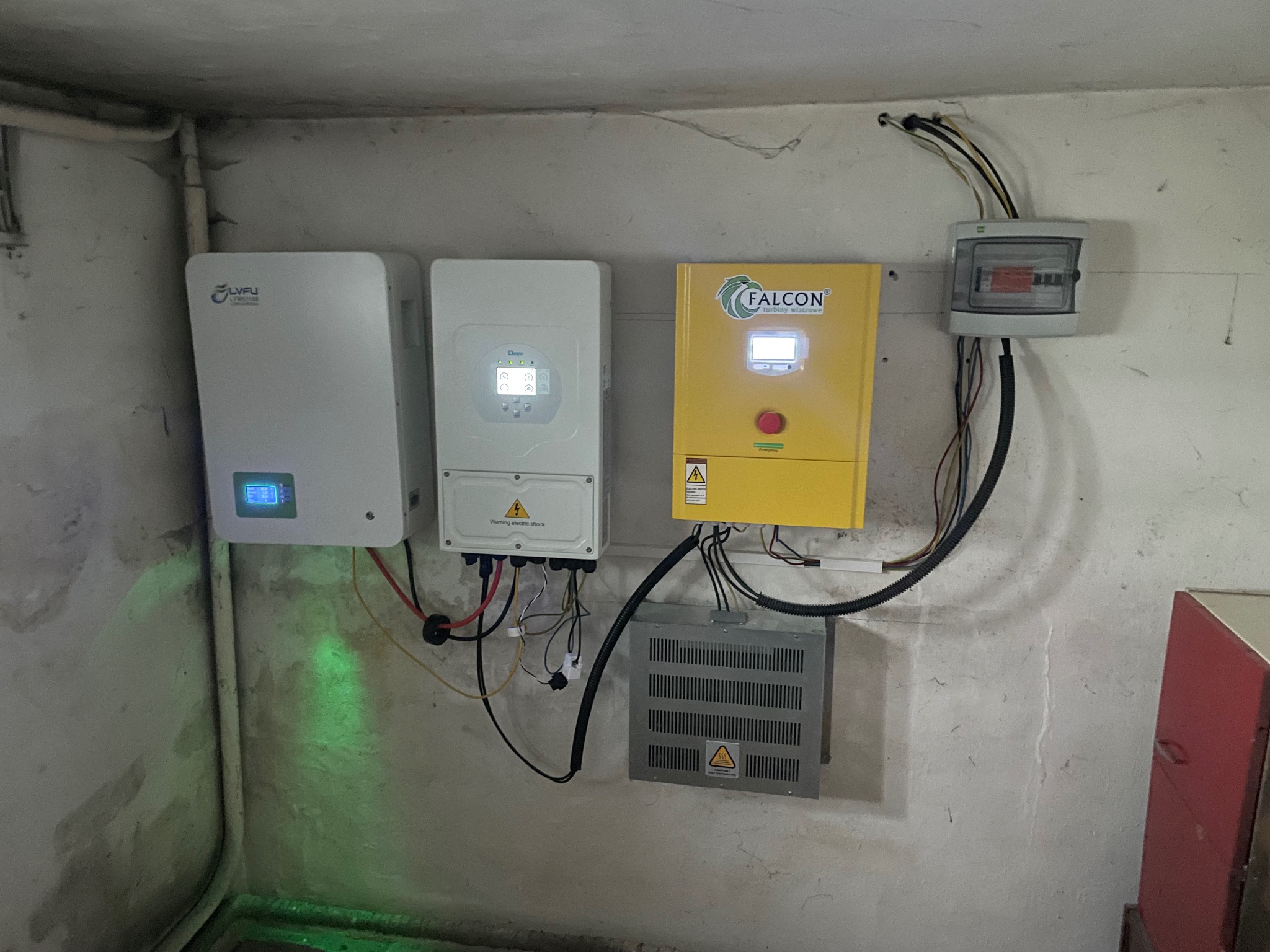 Instalacja fotowoltaiczna w piwnicy: falowniki LVFU, Deye i Falcon turbiny wiatrowe, zabezpieczenia elektryczne. Widoczne okablowanie i elementy montażowe.