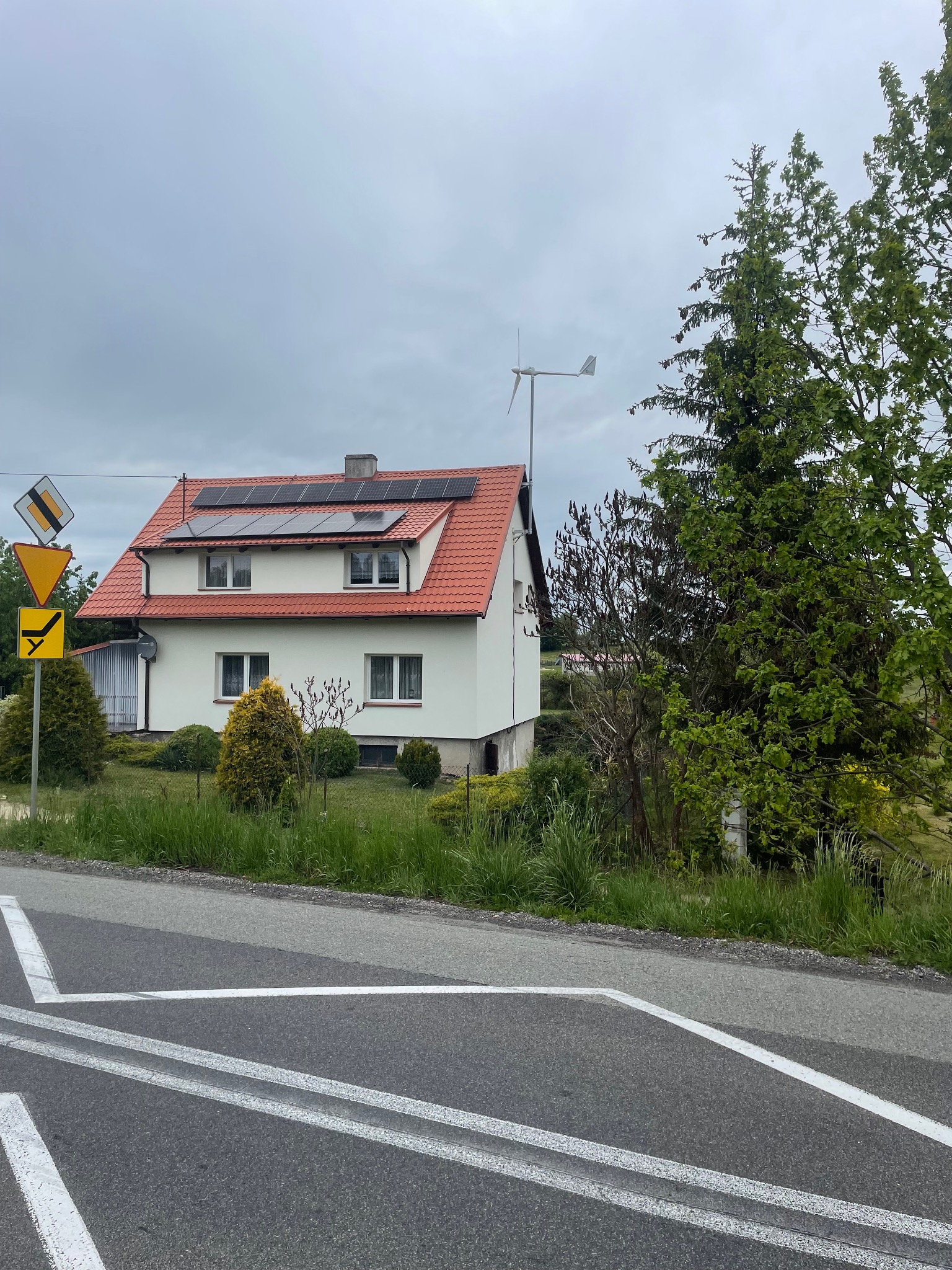 Dom z panelami słonecznymi i małą turbiną wiatrową na dachu, widziany z ulicy z znakami drogowymi. Biała elewacja i czerwona dachówka.