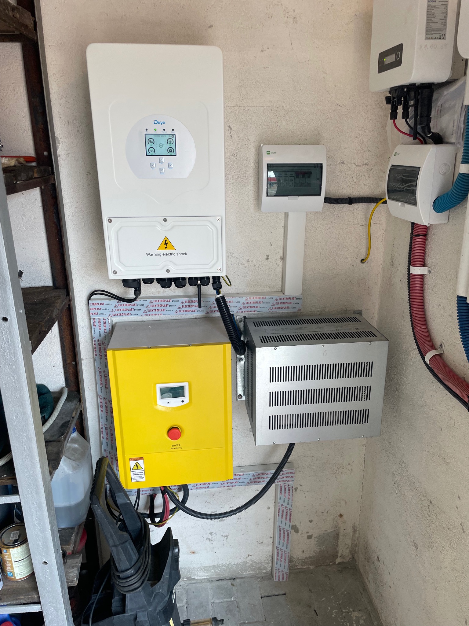 Kompleksowa instalacja fotowoltaiczna w pomieszczeniu: inwerter Deye, żółty transformator, rozdzielnica i okablowanie na ścianie. Widoczne oznaczenia ostrzegawcze.