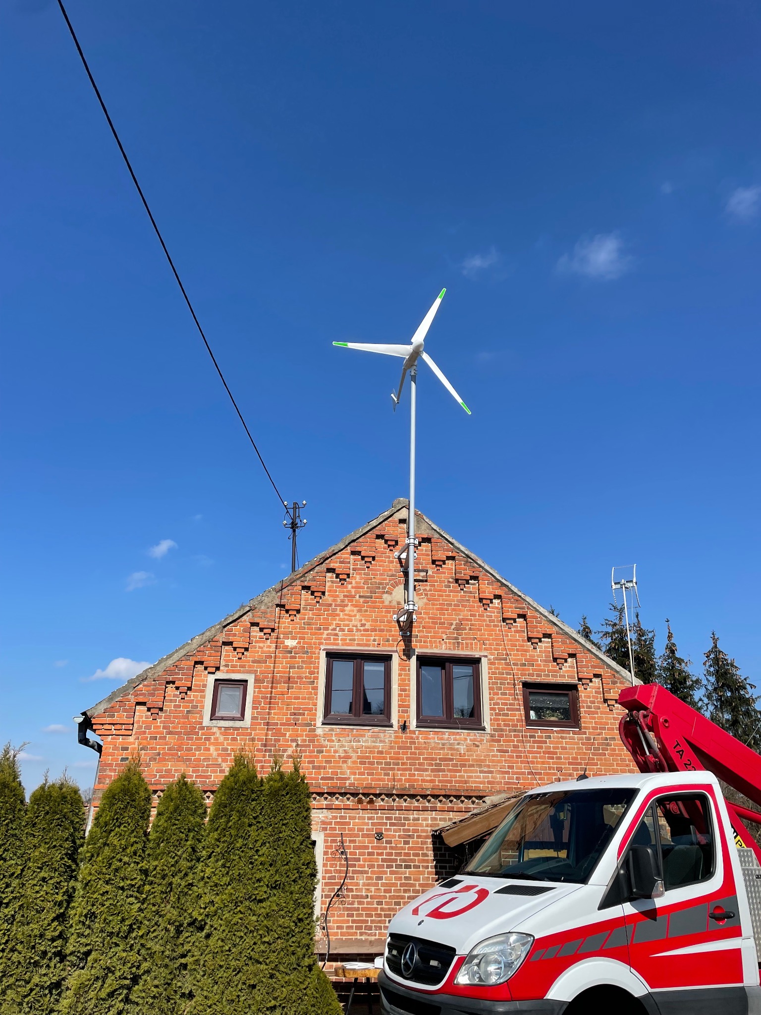 Turbina wiatrowa zamontowana na dachu ceglanego domu, widoczna z poziomu pojazdu serwisowego z wysięgnikiem na tle błękitnego nieba i zielonych tuj.