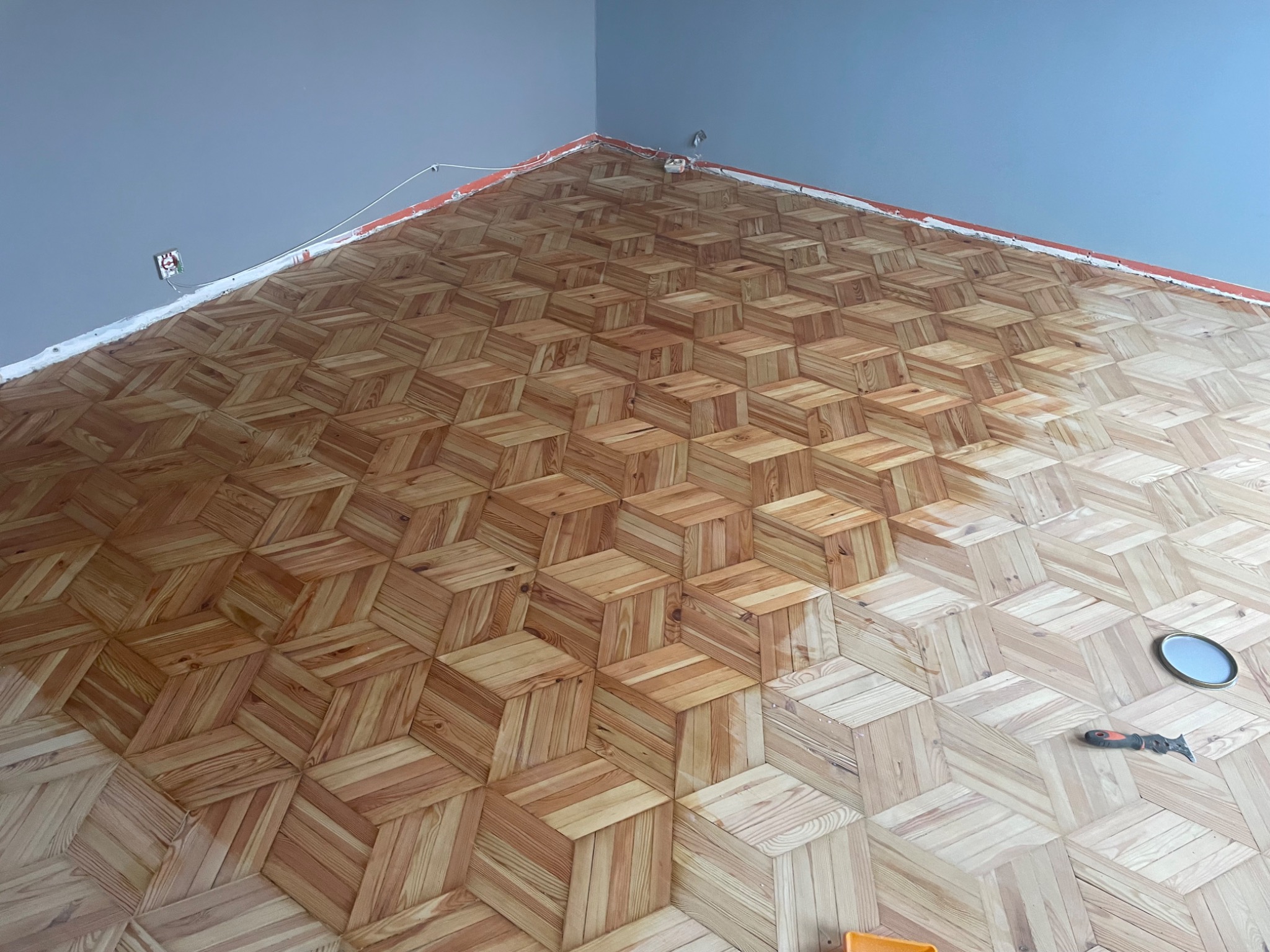 Ukończona podłoga drewniana z wzorem 3D ułożona w rogu pomieszczenia. Widoczne narzędzie i puszka farby. Nietypowy kąt ujęcia podkreśla perspektywę podłogi.