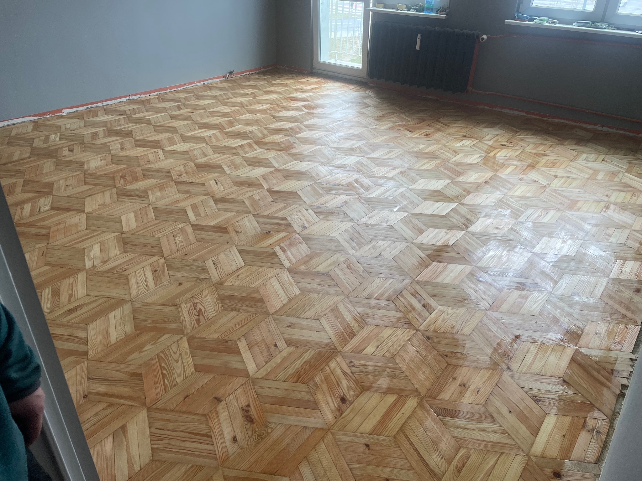 Ukończona podłoga parkietowa z wzorem 3D w jasnym odcieniu drewna, świeżo po lakierowaniu, widok na całe pomieszczenie z grzejnikiem i oknem.