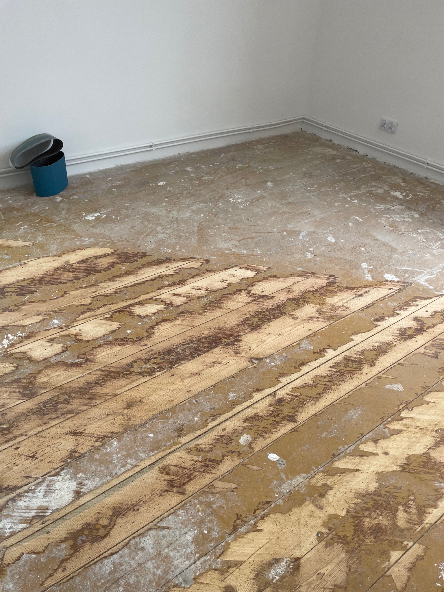 Częściowo cyklinowana podłoga drewniana w jasnym pomieszczeniu. Widoczne deski z resztkami starej powłoki i pyłu. W rogu niebieski pojemnik z pokrywką.