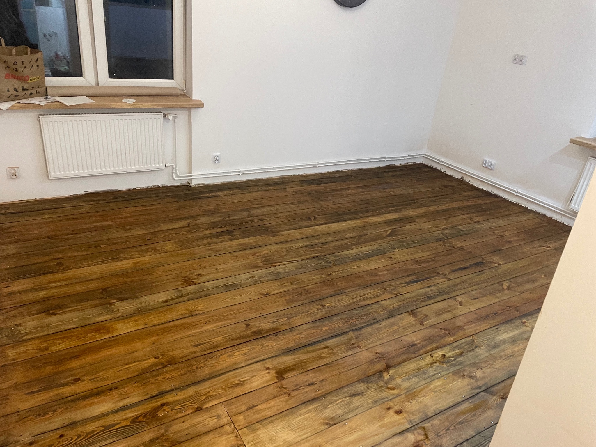 Lakierowana, drewniana podłoga po cyklinowaniu w jasnym pomieszczeniu z białymi ścianami, grzejnikiem i oknem. Widoczne ślady po remoncie przy listwach przypodłogowych.