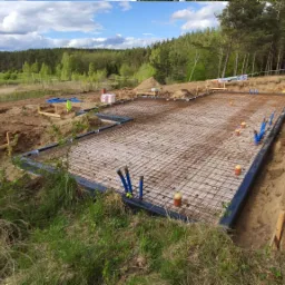 Zbrojenie fundamentu płyty w gruncie, widoczne niebieskie rury instalacyjne, szalunek obłożony folią, teren budowy zalesiony w tle.