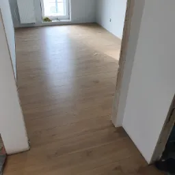 Nowo położone panele podłogowe w jasnym odcieniu drewna, widoczne z perspektywy wejścia do pokoju z niedokończonymi obramowaniami drzwi, w tle grzejnik pod oknem.