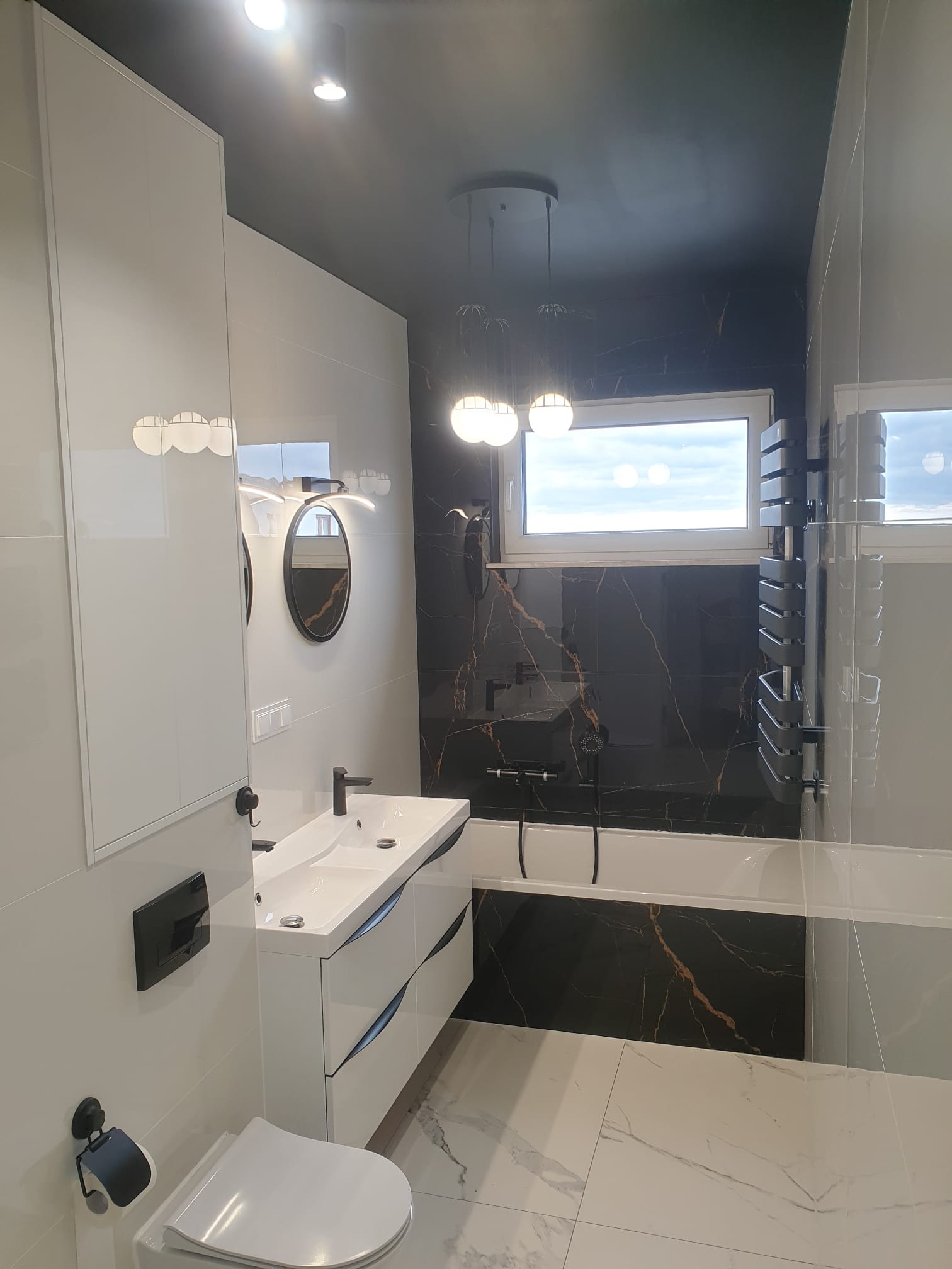 Nowoczesna łazienka z czarno-białymi płytkami imitującymi marmur, wisząca szafka z umywalką, okrągłe lustro i dekoracyjne lampy wiszące.