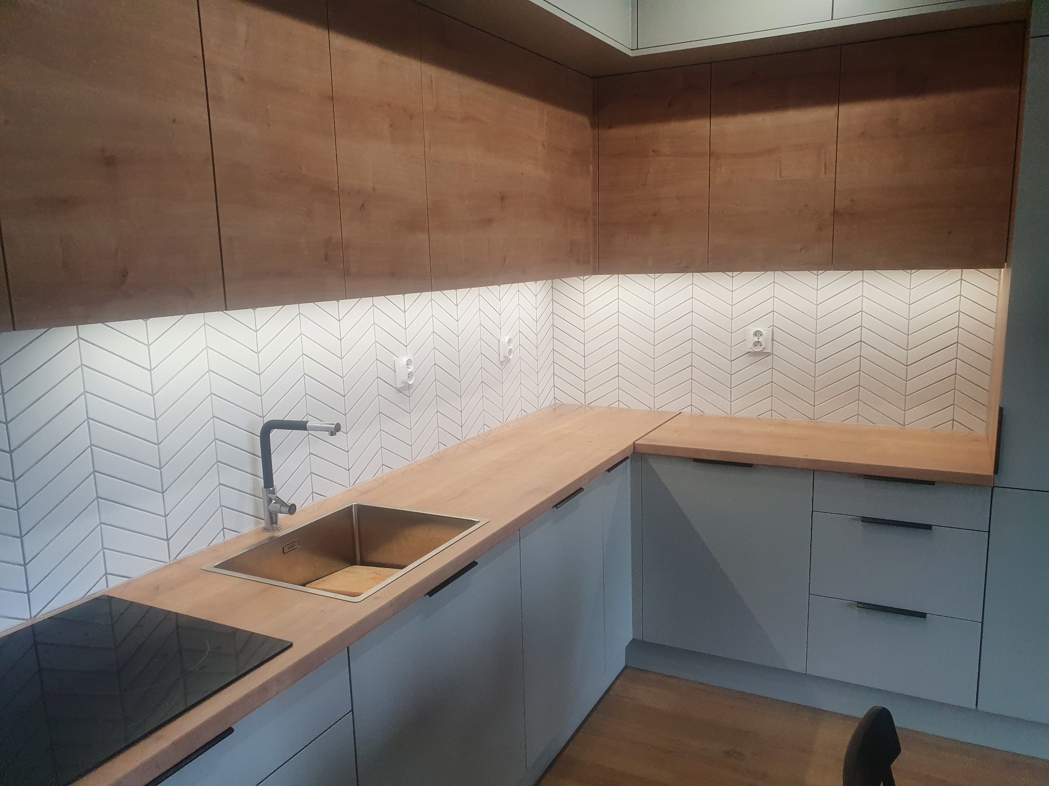 Nowoczesna kuchnia z drewnianymi szafkami górnymi, szarymi szafkami dolnymi, blatem z drewna i białymi płytkami ułożonymi w jodełkę nad blatem.