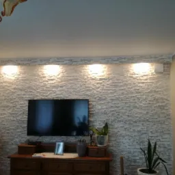 Ściana z dekoracyjnej, jasnej cegły z podświetleniem LED, na której wisi telewizor. Poniżej drewniana komoda z dekoracjami i roślinami.