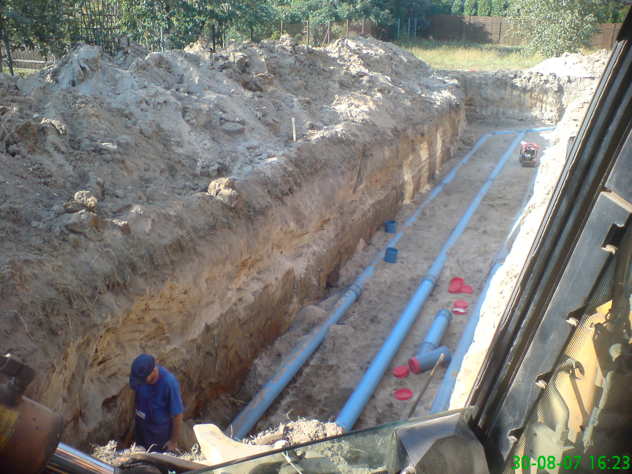 Głęboki wykop w Gliwicach z ułożonymi rurami kanalizacyjnymi, widok z kabiny koparki na pracownika w niebieskiej koszuli i czapce, kontrolującego instalację.