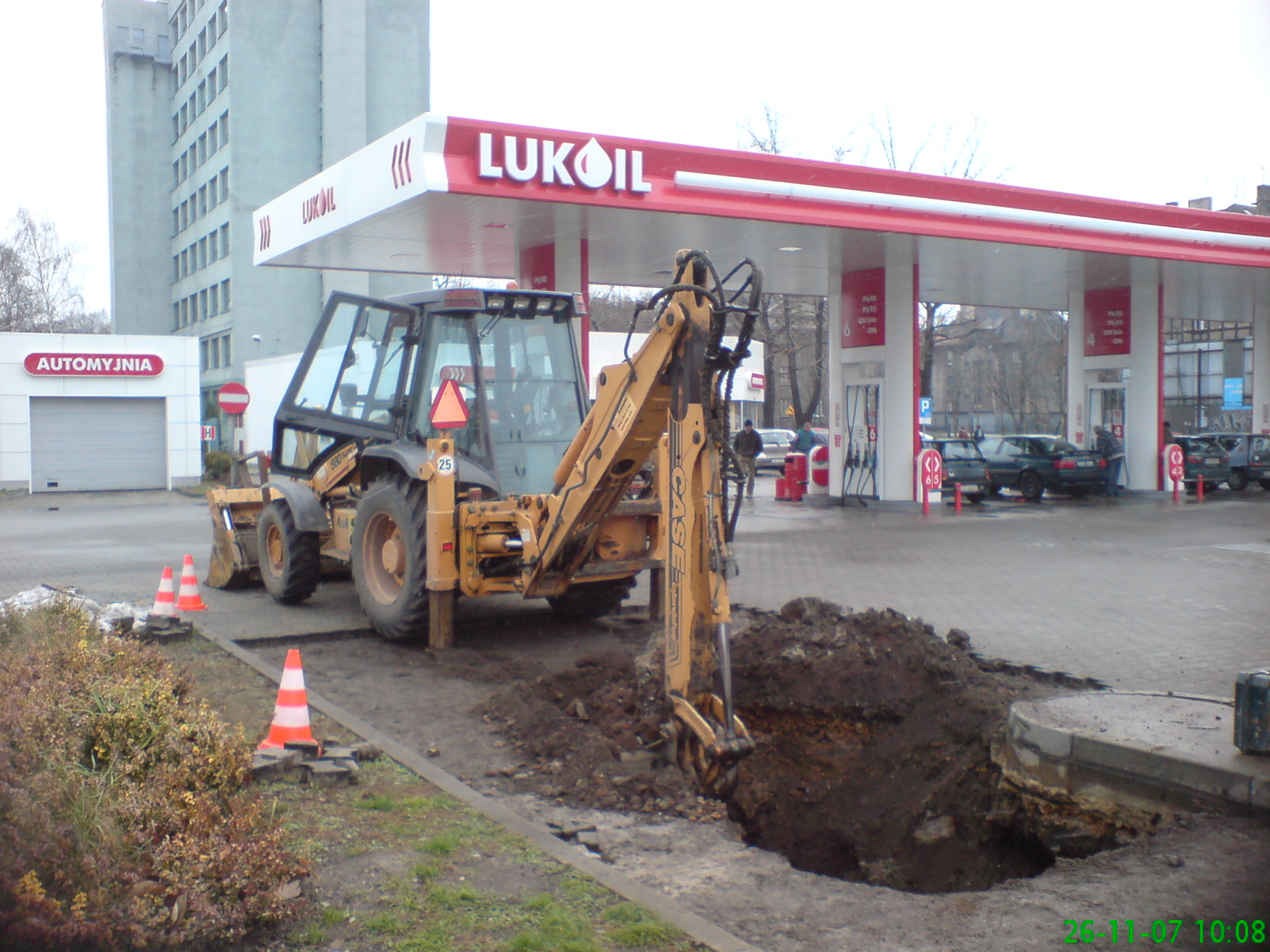 Żółta koparko-ładowarka CASE 580ST z wysuniętym ramieniem kopiąca dół w ziemi przy stacji benzynowej Lukoil, zabezpieczona pomarańczowymi pachołkami.