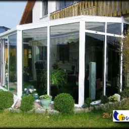 Ogród zimowy z białymi ramami okiennymi w Letnicy, otoczony niskimi krzewami i rabatami kwiatowymi, widok z zewnątrz na przeszkloną konstrukcję przylegającą do domu.