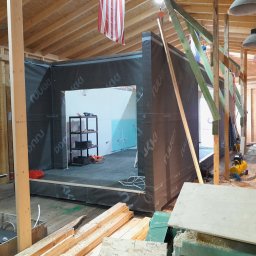 MTS SCHREINER - Wnętrze w trakcie budowy domu szkieletowego w Tarnowie Podgórnym. Widoczne drewniane elementy konstrukcyjne i czarna membrana. W tle regał i narzędzia.