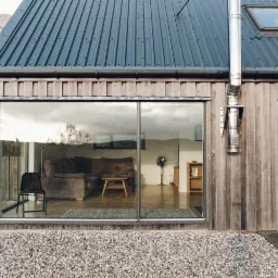 Nowoczesny dom z drewnianą elewacją, dużymi oknami i metalowym dachem, widok z zewnątrz na salon z kanapą, krzesłem i stołem oraz jadalnię z drewnianym stołem i krzesłami.