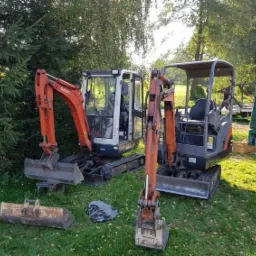Dwa mini-koparki Kubota stoją na trawie obok siebie, z dodatkową łyżką leżącą na ziemi i przyczepą w tle.