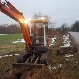 Pomarańczowa mini koparka Kubota z włączonymi światłami roboczymi, pracująca na poboczu drogi, z widoczną wykopaną ziemią przed łyżką i resztkami śniegu na trawie.