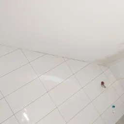 Ściana wyłożona białymi, błyszczącymi płytkami ułożonymi pod kątem, widoczne gniazda elektryczne bez osprzętu, fragment sufitu z widocznymi śladami po gładzi.