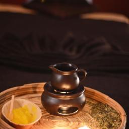 Sabai Spa - Przygotowany zestaw do aromaterapii na wiklinowej tacy: ceramiczny podgrzewacz z olejkiem, miseczka z żółtym proszkiem i zioła, oświetlone dwoma tealightami, w tle łóżko do zabiegów.