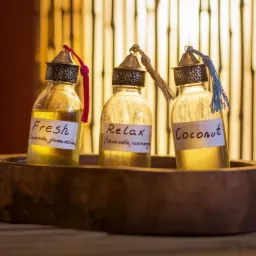 Trzy szklane buteleczki z olejkami eterycznymi (Fresh, Relax, Coconut) z dekoracyjnymi korkami i etykietami, umieszczone w drewnianej tacy na tle bambusowej ściany.