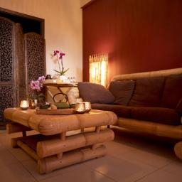 Sabai Spa - Stylowe wnętrze z bambusowymi meblami: sofa z brązowymi poduszkami, stolik z herbatą i lampionami, ozdobny parawan i kwiaty, tworzące relaksującą atmosferę.