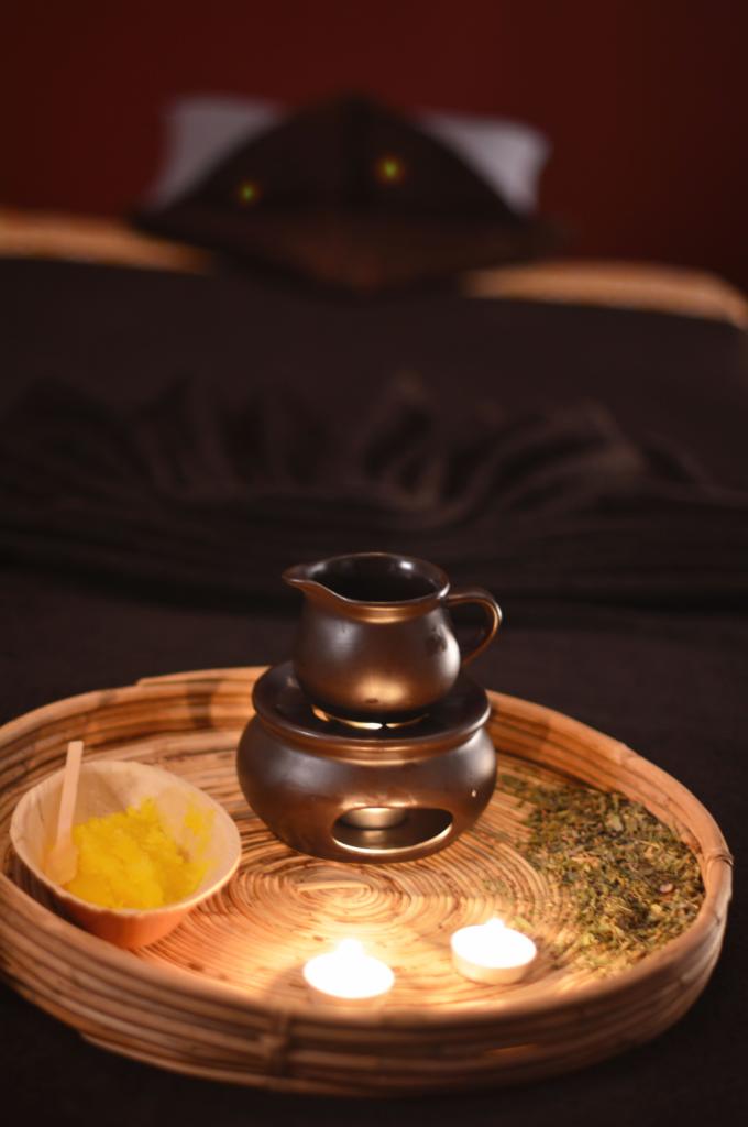 Przygotowany zestaw do aromaterapii na wiklinowej tacy: ceramiczny podgrzewacz z olejkiem, miseczka z żółtym proszkiem i zioła, oświetlone dwoma tealightami, w tle łóżko do zabiegów.