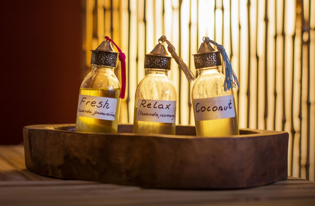 Trzy szklane buteleczki z olejkami eterycznymi (Fresh, Relax, Coconut) z dekoracyjnymi korkami i etykietami, umieszczone w drewnianej tacy na tle bambusowej ściany.