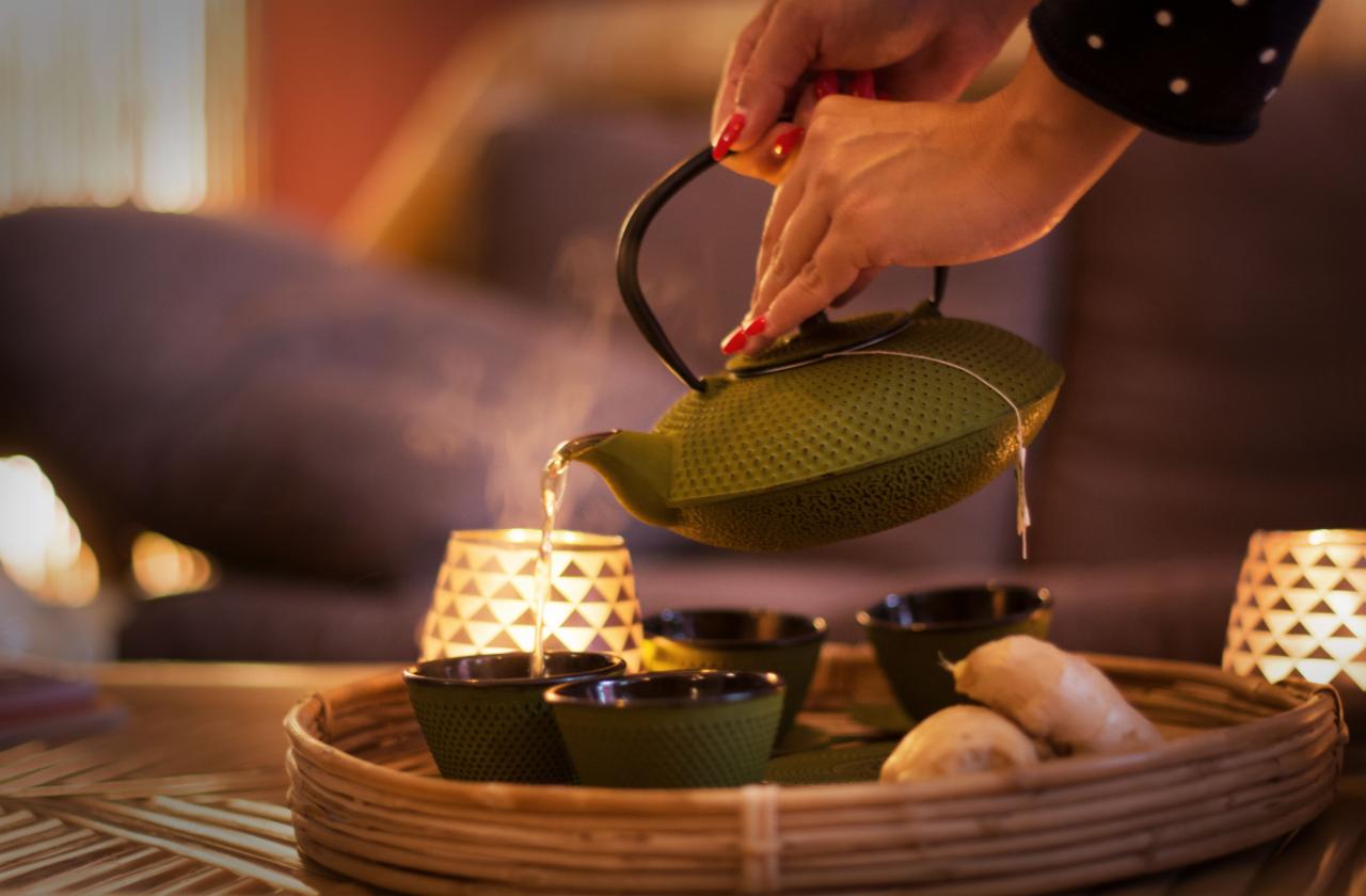 Osoba z czerwonym manicure nalewa parujący napar z oliwkowego, żeliwnego czajnika do pasujących czarek na bambusowej tacy obok kawałków imbiru, w tle rozproszone światło świec i miękka sofa.
