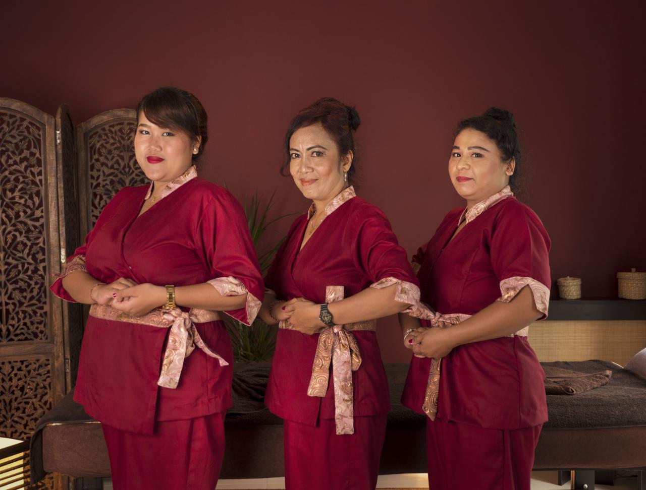 Trzy kobiety w bordowych strojach spa z ozdobnymi pasami, stojące przed łóżkiem do masażu i dekoracyjnym parawanem, prezentujące ofertę hotelu SPA.