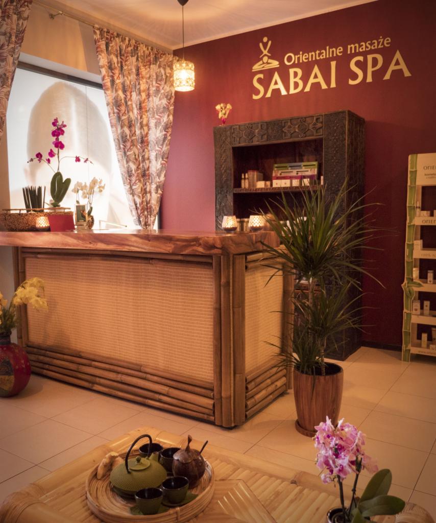 Recepcja w orientalnym SPA z bambusowym blatem, widoczne logo 'Sabai SPA' na ścianie, dekoracje roślinne i zestaw do herbaty na stoliku.